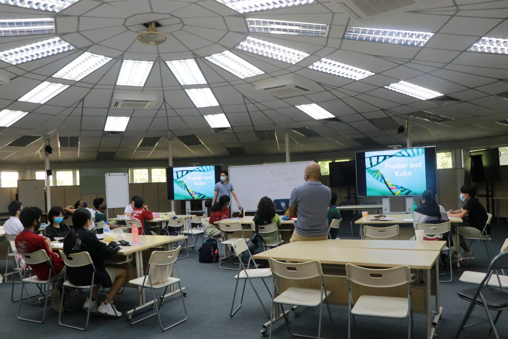 Students wearing masks sit at tables in a domed classroom while a teacher presents a "Popper and Kuhn" slideshow.
